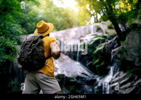 Vista posteriore dell'irriconoscibile escursionista maschile in piedi sul masso e ammirando la cascata nella foresta Foto Stock