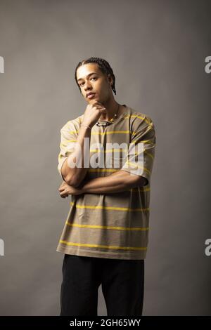 Giovane modello maschile afro-americano con capelli intrecciati vestito con camicia oversize a righe e collana che guarda la fotocamera contro il backgrou grigio Foto Stock