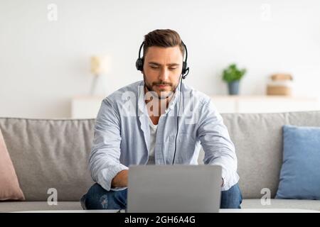 Servizio di assistenza hotline. Operatore di contact centre maschile millenario in cuffie che lavora con il computer portatile a casa in salotto Foto Stock
