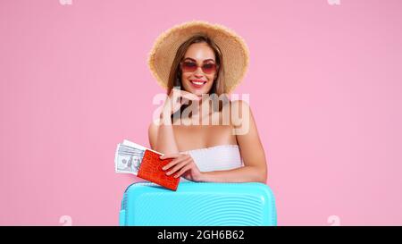 Ragazza giovane positiva in cappello di paglia e occhiali da sole con biglietti e Suitcase Pink Studio sfondo, sorrisi guardando la macchina fotografica Foto Stock