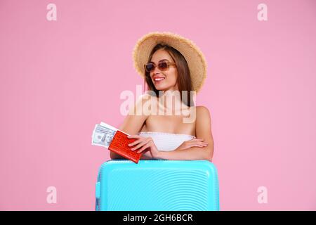 Ragazza giovane positiva in cappello di paglia e occhiali da sole con biglietti e Suitcase Pink Studio sfondo, sorrisi guardando via Foto Stock