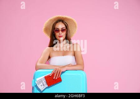 Ragazza giovane positiva in cappello di paglia e occhiali da sole con biglietti e Suitcase Pink Studio sfondo guardando la fotocamera Foto Stock