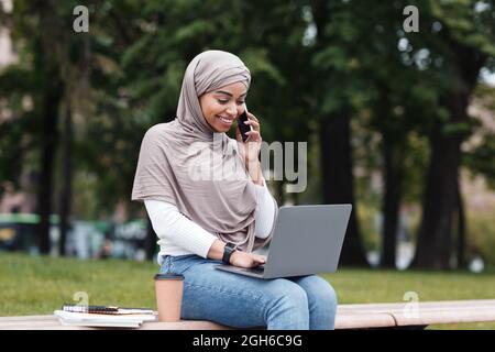 Allegro bella donna d'affari nero lavora all'aperto con caffè e laptop, chiamate per telefono con il cliente in parco urbano Foto Stock