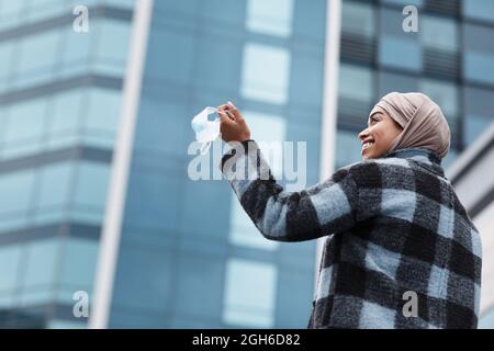 Felice sorridente bella donna nera in hijab e cappotto tiene in mano la maschera medica e gode di libertà all'aperto Foto Stock