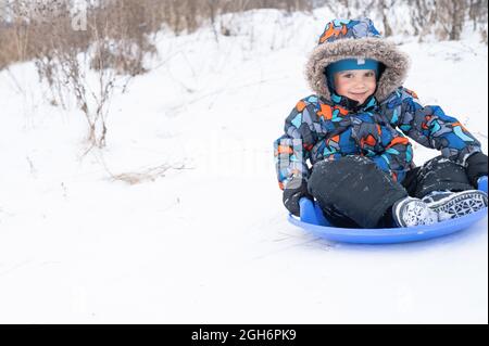 un ragazzo felice carino di cinque anni è attività e divertimento trascorrere le sue vacanze invernali nevose cavalcando uno scivolo su una slitta Foto Stock