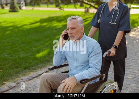 Uomo anziano, ricoverando il paziente in sedia a rotelle che parla al telefono mentre cammina con l'infermiere nel parco in una giornata estiva Foto Stock