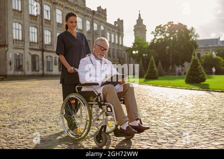 Shot completo di medico anziano handicappato in sedia a rotelle indossando camice da laboratorio e occhiali con giovane infermiere che lo assiste all'aperto Foto Stock