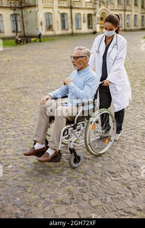 Scatto a tutta lunghezza di cura giovane infermiere in maschera protettiva prendersi cura di uomo anziano handicappato paziente in sedia a rotelle durante una passeggiata all'aperto Foto Stock