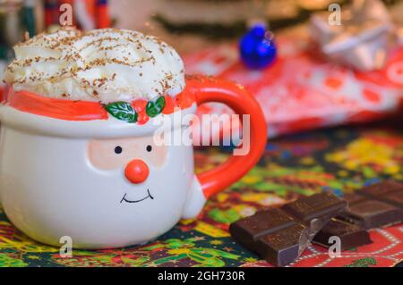 Cioccolata calda di Natale in tazza Babbo Natale con panna montata e cioccolato rasato con quadrati di cioccolato sul lato. Bevanda calda di Natale. Natale p Foto Stock