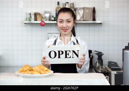 Il giovane negoziante asiatico con un sorriso tiene un cartello APERTO di fronte al banco di una caffetteria. Atmosfera mattutina in una caffetteria. Foto Stock