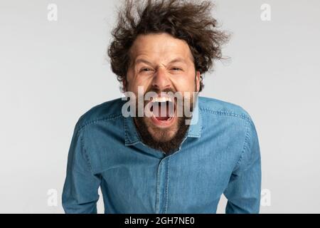 uomo arrabbiato non rasato con capelli ricci in denim camicia urla e furioso su sfondo grigio in studio Foto Stock