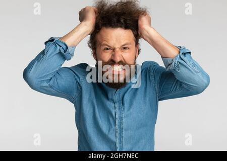 l'uomo arrabbiato in camicia di denim essendo nervoso e tirando i capelli fuori, facendo i volti sconvolti e grimacing su sfondo grigio in studio Foto Stock