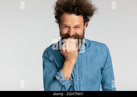 divertente modello casual in camicia in denim con barba lunga e capelli ricci che tengono le dita alla bocca, mordendo e facendo facce sciocche su sfondo grigio in studio Foto Stock