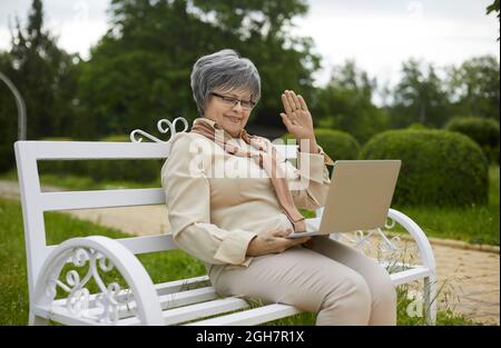 Donna anziana felice seduta sulla panca del parco, videochiamata famiglia e hello onding allo schermo del laptop Foto Stock