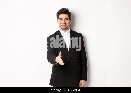 Ritratto di bell'uomo d'affari in abito nero, che si estende la mano per la stretta di mano, salutare i partner commerciali e sorridere, benvenuto in azienda, in piedi Foto Stock