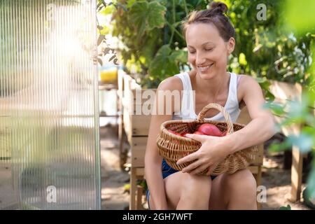 Ritratto di giovane adulto bella femmina agricoltore seduta contro serra raccolta fresco pomodori rossi maturi in vimini basket casa giardino letto Foto Stock