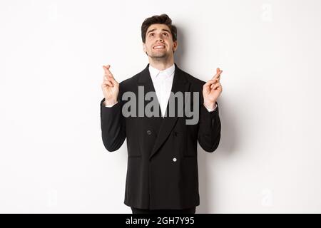 Ritratto di bell'uomo d'affari teso e preoccupato, incrociando le dita e guardando in su, pregando dio, facendo un desiderio, in piedi contro sfondo bianco in Foto Stock