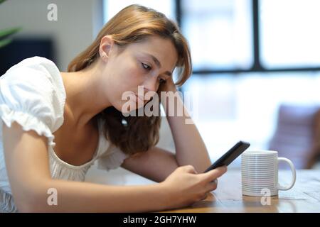 Donna frustrata e annoiata utilizzando smartphone controllare contenuti seduti a casa Foto Stock