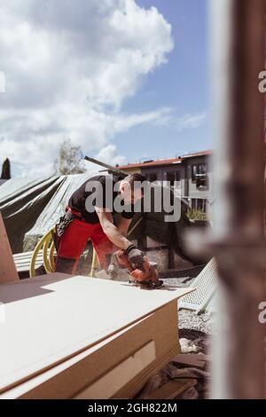 Carpentiere maschio che taglia la tavola di legno dalla sega elettrica al luogo di costruzione durante la giornata di sole Foto Stock