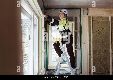 L'intera lunghezza del telaio della finestra di misurazione dell'appaltatore dell'edificio femmina è in piedi sulla scala a gradini in cantiere Foto Stock