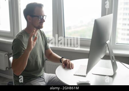 Giovane allegro che indossa occhiali eleganti e abbigliamento informale utilizzando l'app per computer per la connessione video, uomo libero che accoglie i partecipanti online, ondeggiandosi in webcam. Concetto di videochiamata Foto Stock