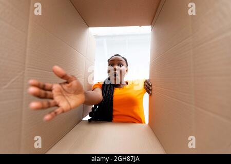 Vista dal basso della donna nera che riceve un ordine online, disimballando il pacco in cartone al chiuso Foto Stock
