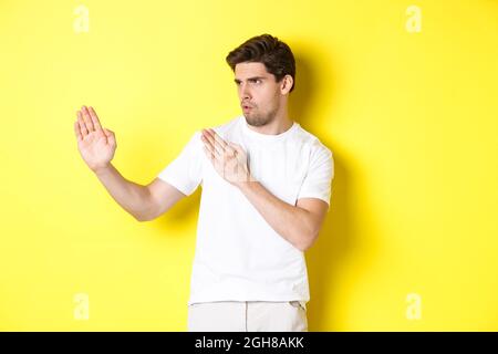 Uomo che mostra abilità kung-fu, arti marziali movimento ninja, in piedi in bianco t-shirt pronto a combattere, in piedi su sfondo giallo Foto Stock