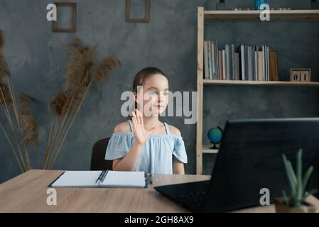 Piccola ragazza caucasica sorridente e agitando la mano mentre si siede a casa al tavolo con un computer portatile. Comunicazione online, formazione a distanza. Copia spac Foto Stock