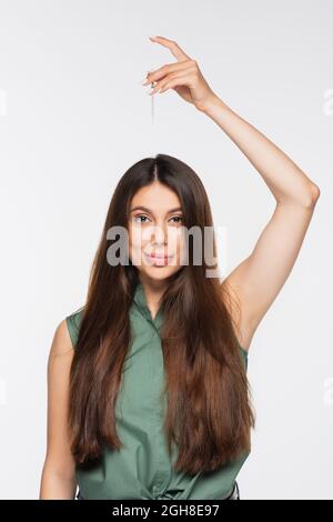 giovane donna allegra con capelli lucidi applicando siero di trattamento sulla parte superiore della testa isolato su grigio Foto Stock