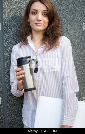 Ritratto femminile di giovane donna con computer portatile in argento e tazza di caffè in attesa di un incontro vicino al muro grigio scuro al mattino, lavoro remoto, coffee break Foto Stock