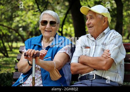 Coppia anziana felice seduta su una panca. Uomo anziano e donna in occhiali da sole ridendo insieme, tempo libero nel parco, vita in pensione Foto Stock