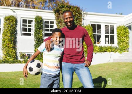 Ritratto di sorridente padre afroamericano con figlio che abbraccia e tiene il calcio in giardino soleggiato Foto Stock