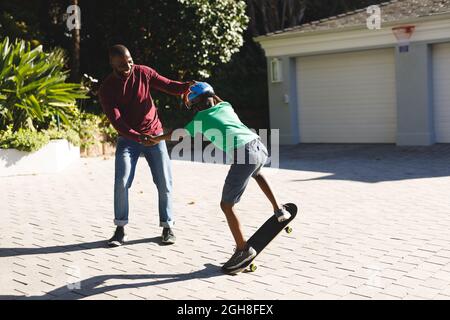 Il padre afroamericano sorride e aiuta il figlio a bilanciarsi sullo skateboard in giardino Foto Stock