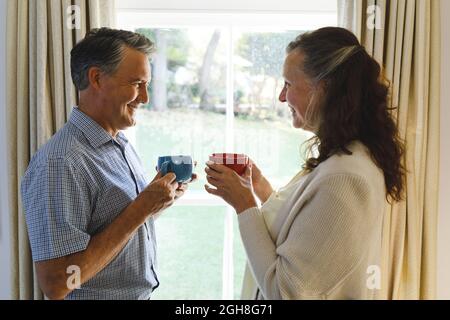Felice coppia caucasica senior in piedi accanto alla finestra, tenere tazze. Stile di vita pensione, trascorrere il tempo a casa. Foto Stock