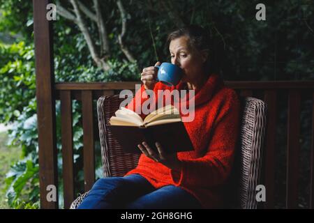 Felice anziana donna caucasica seduta su sedia, libro di lettura e bere caffè in giardino Foto Stock