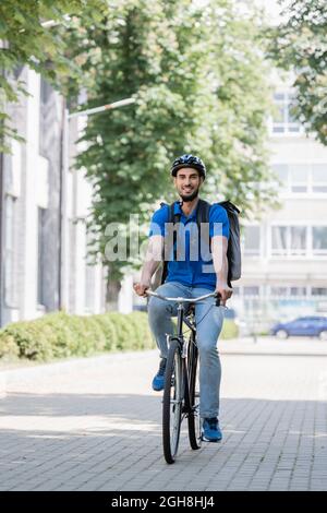 Buon corriere arabo in casco di sicurezza e zaino in bicicletta su strada urbana Foto Stock