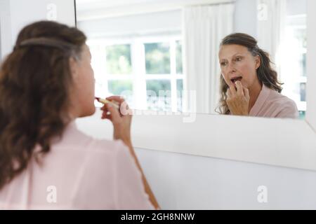 Donna caucasica anziana felice in bagno, guardando nello specchio, applicando rossetto Foto Stock