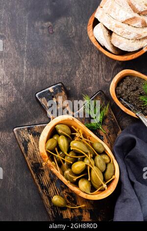 Capperi in scatola in piatto di legno, tapenade e pane su un vecchio sfondo alla moda di legno scuro. Vista dall'alto Foto Stock
