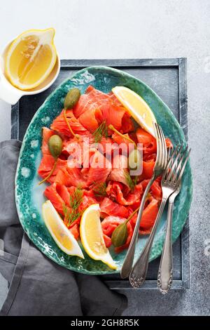 Fette di salmone leggermente salato con capperi, limone e aneto su un piatto di ceramica su sfondo grigio di cemento. Vista dall'alto Foto Stock