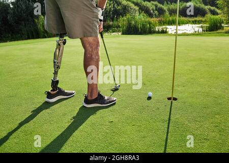 Golfer professionista con gamba protesica che colpisce con putter sulla sfera di golf durante il golf alla giornata di sole con le ombre lunghe Foto Stock