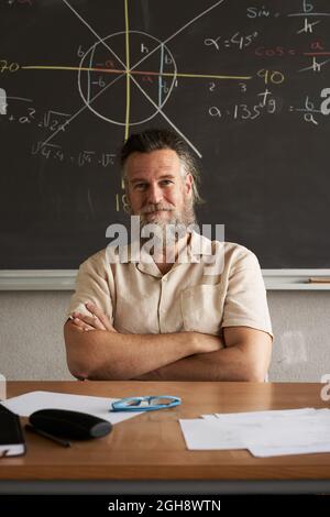 Foto verticale. L'insegnante allegro guarda la macchina fotografica con le braccia incrociate. Classe matematica. Foto Stock