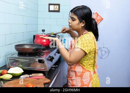 Una giovane donna indiana bella che indossa grembiule cucina e degustazione di cibo in cucina domestica su stufa a gas Foto Stock