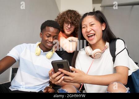 Gruppo di studenti adolescenti di diverse etnie che utilizzano i telefoni cellulari. Ragazza cinese che guarda la macchina fotografica mentre ride. Foto Stock