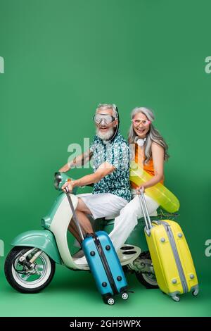 coppia multietnica e felice di mezza età in occhiali che tengono il bagaglio mentre si guida ciclomotore sul verde Foto Stock