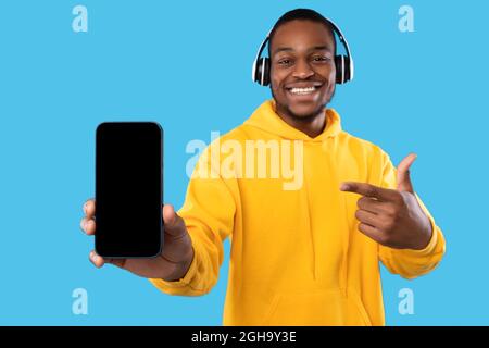 Uomo nero che mostra il telefono schermo vuoto indossando cuffie, sfondo blu Foto Stock