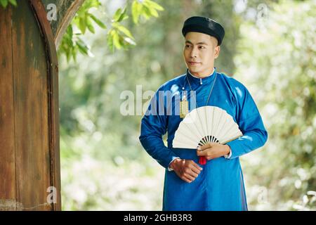 Ritratto di bel giovane vietnamita in tradizionale ao dai vestito a piedi nel parco con ventilatore in legno in mani Foto Stock