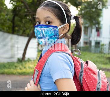 Primo piano di una ragazza indiana felice studente bambino in uniforme blu scuola con sacco rosso e maschera naso protezione andare a scuola Foto Stock