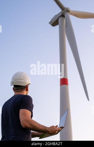 L'ingegnere maschile nel casco controlla e mantiene il funzionamento di una turbina eolica utilizzando un computer portatile sullo sfondo del mulino a vento Foto Stock