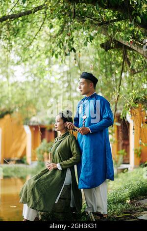 Bella appena sposata vietnamita coppia in ao dai vestiti seduti da stagno e guardando l'acqua Foto Stock