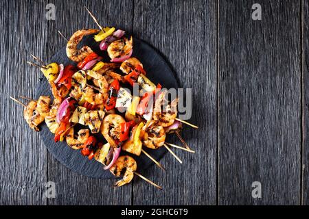 Spiedini di gamberetti alla griglia con cipolla rossa, pepe dolce e ananas su un piatto di pietra nera, piatto, spazio libero Foto Stock